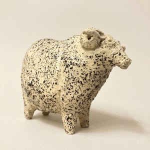 May include: A ceramic sheep figurine with a speckled cream and black pattern. The sheep has curled horns and is standing on four legs. The figurine is a decorative item.