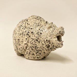Könnte beinhalten: Keramik-Bärenfigur mit gesprenkeltem, cremefarbenem Körper und dunkelbraunen Flecken. Der Bär ist mit offenem Maul modelliert, die Zähne sind sichtbar. Die Figur ist ein dekoratives Objekt, wahrscheinlich zur Ausstellung.