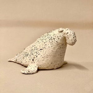 May include: A ceramic sculpture of a seal, painted white with black speckles. The seal is in a seated position, with its flippers extended and its head tilted upwards. The sculpture has a textured surface.