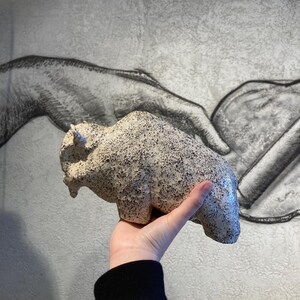 May include: A speckled, off-white ceramic bison sculpture. The bison is held in a hand, showcasing its textured surface and detailed form. The sculpture has a small tail and horns. The background features a gray and white drawing of hands.