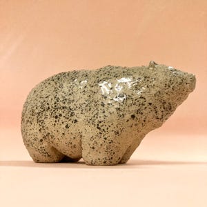 Op de afbeelding: Een keramieken berenbeeldje in een staande pose. De beer is lichtbeige met zwarte spikkels en een glanzende afwerking. Het beeldje heeft een afgerond lichaam en staat tegen een perzikkleurige achtergrond.