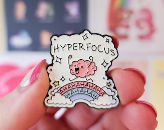 Op de afbeelding: Witte emaille pin met de woorden "HYPERFOCUS" boven een roze hersenen met een lachend gezicht. De pin heeft een regenboog en het woord "AHAHAHA" in een herhalend patroon. De pin wordt door een persoon vastgehouden.