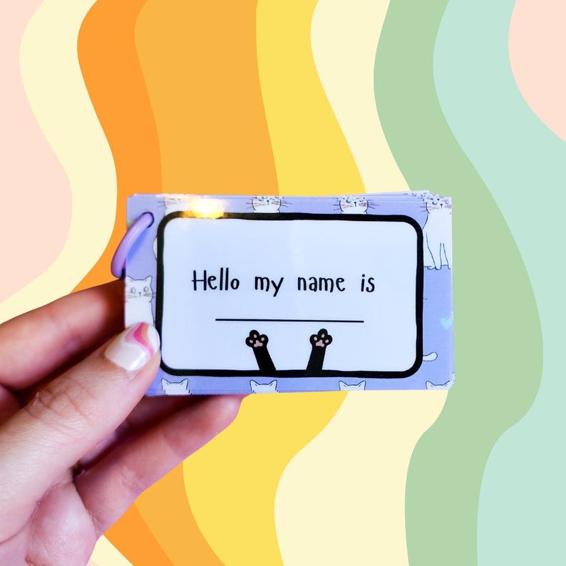 May include: A purple and white name tag with a black cat paw print design. The tag says "Hello my name is" with a blank line for writing.