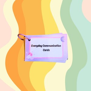 May include: A stack of Everyday Communication Cards with a purple border and a silver ring. The cards have a white background with the text "Everyday Communication Cards" in black. The background is a colorful, wavy pattern.