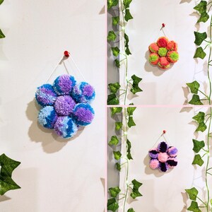 May include: Three wall hangings in the shape of flowers made of pom poms. The first flower is purple and blue, the second is orange and green, and the third is pink and purple.