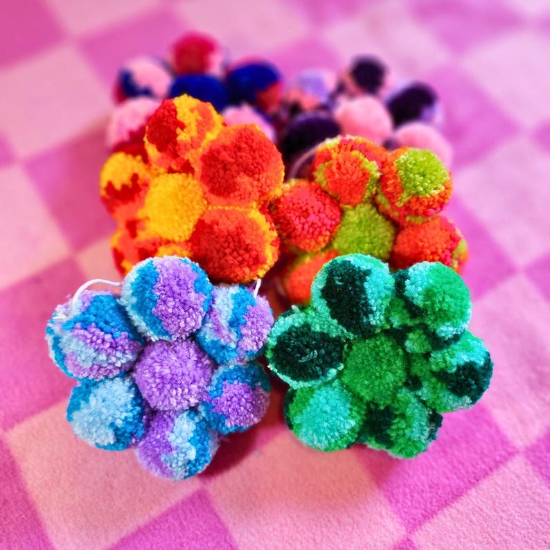 May include: A set of five colorful pom-pom flowers, each with a different color scheme. The flowers are made of yarn and have a fluffy texture. They are arranged in a cluster on a pink and white checkered background.