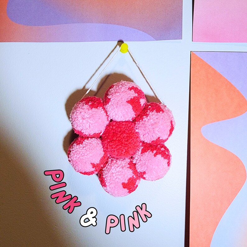 May include: A flower-shaped wall hanging made of pink and red pom-poms. The pom-poms are attached to a string and suspended from a yellow push pin. The text "PINK & PINK" is in the lower left corner.