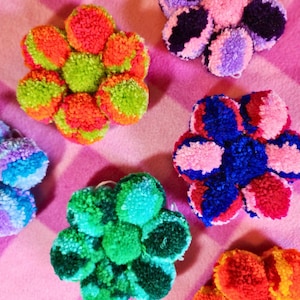 May include: Six colorful pom-pom flowers in various colors, including pink, blue, green, and orange. The flowers are arranged on a pink and white striped background.