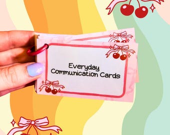 May include: A set of pink and white communication cards with the text "Everyday Communication Cards" on the front. The cards are decorated with a cherry and bow design.
