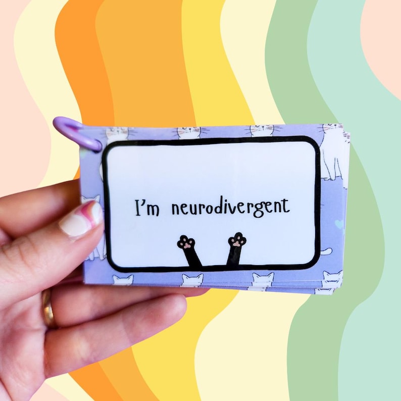 May include: A purple card with white cats printed on it. The card has a black border and says "I'm neurodivergent" in black text. Two black cat paws are sticking out from the bottom of the card.