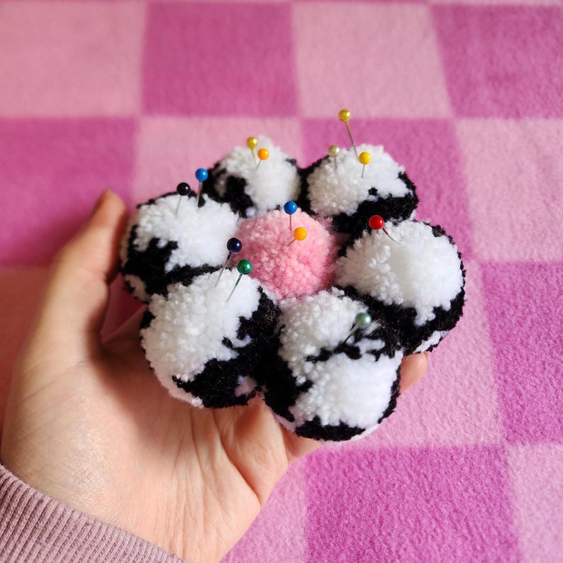 May include: A handmade pin cushion shaped like a flower, featuring black and white pom-poms around a pink center. Colorful pins are inserted into the pom-poms. The cushion is held in a hand against a pink checkered background.