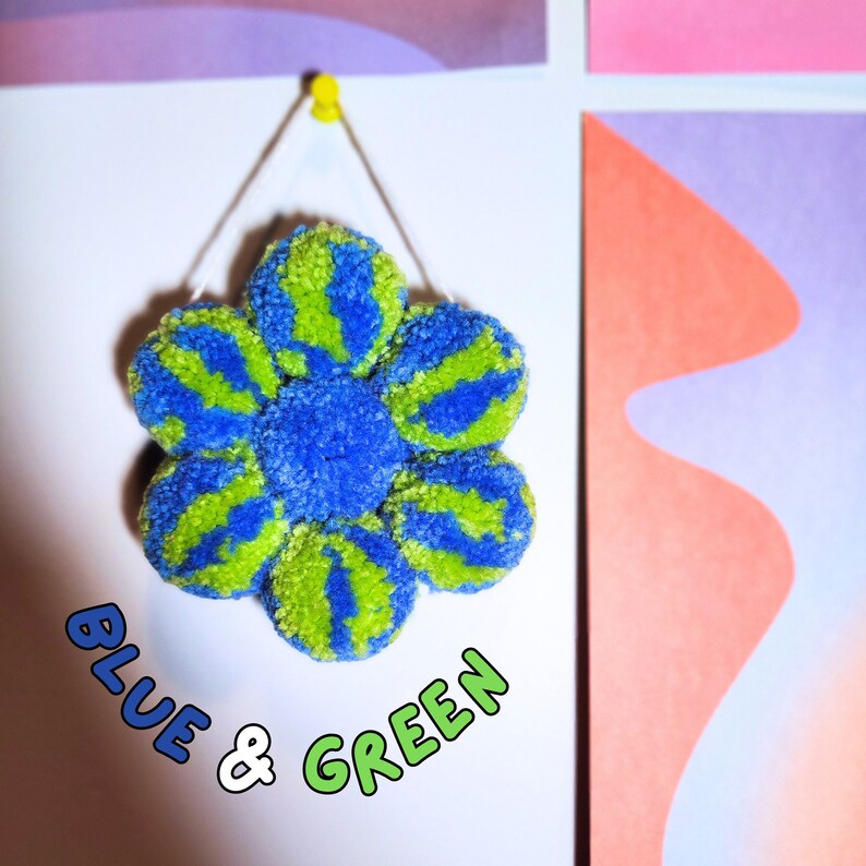 May include: A handmade, blue and green pom-pom flower decoration hanging on a white wall. The flower has a blue center and alternating blue and green petals. The text "BLUE & GREEN" is written below the flower.