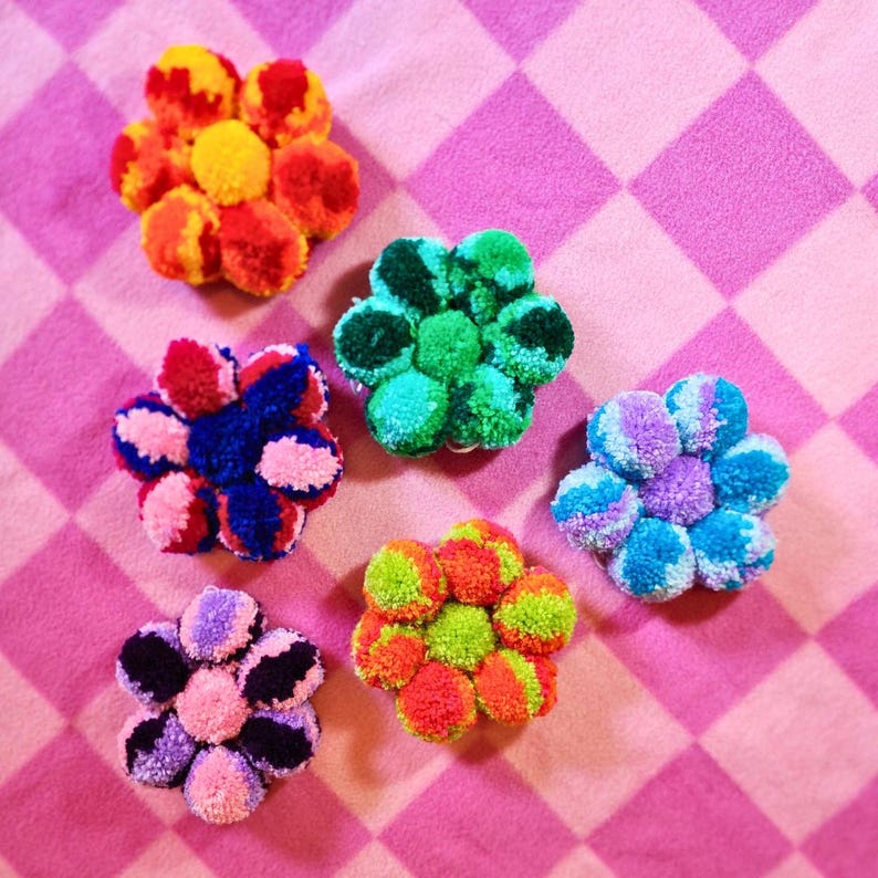 May include: Six colorful pom-pom flowers in various colors, including red, orange, yellow, green, blue, and purple, arranged on a pink and white checkered background.