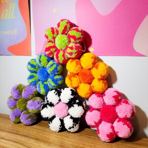 May include: A collection of handmade pom-pom flowers in various colors, including green, blue, orange, purple, pink, and black and white. Each flower is composed of multiple pom-poms arranged in a floral shape, perfect for home decor.