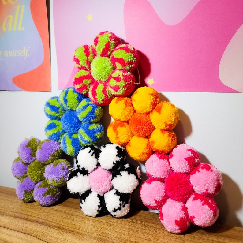 May include: A collection of handmade pom-pom flowers in various colors, including green, blue, orange, purple, pink, and black and white. Each flower is composed of multiple pom-poms arranged in a floral shape, perfect for home decor.