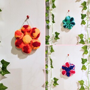May include: Three pom-pom flower wall hangings in different color combinations. The first is red, orange, and yellow. The second is teal and green. The third is pink, blue, and white.