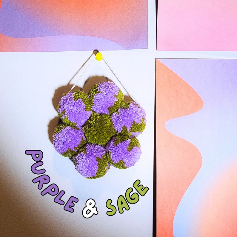 May include: A handmade pom-pom decoration in a flower shape, featuring purple and sage green yarn. The decoration is suspended from a white string and a yellow push pin. The text "PURPLE & SAGE" is displayed in a playful font.