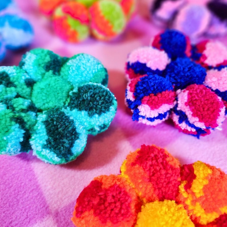May include: A collection of colorful pom-pom flowers in various shades of blue, green, pink, red, and yellow. The flowers are made of yarn and have a fluffy texture.