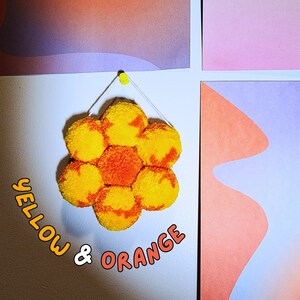 May include: A handmade, flower-shaped wall hanging composed of yellow and orange pom-poms. The text "YELLOW & ORANGE" is written in a playful font. The artwork is suspended from a white wall with a yellow push pin, with colorful abstract art in the background.