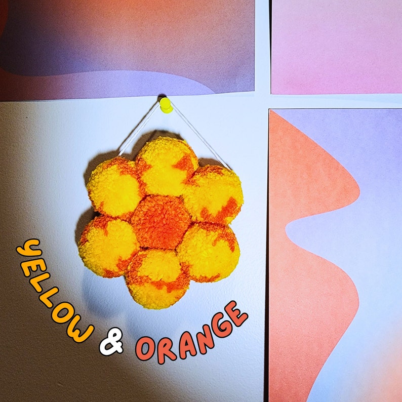 May include: A handmade, flower-shaped wall hanging composed of yellow and orange pom-poms. The text "YELLOW & ORANGE" is written in a playful font. The artwork is suspended from a white wall with a yellow push pin, with colorful abstract art in the background.
