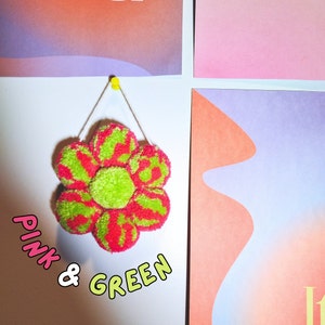 May include: A handmade, flower-shaped wall hanging made of pink and green yarn. The flower is attached to a white wall with a yellow push pin. The text "PINK & GREEN" is written in a playful font below the flower.