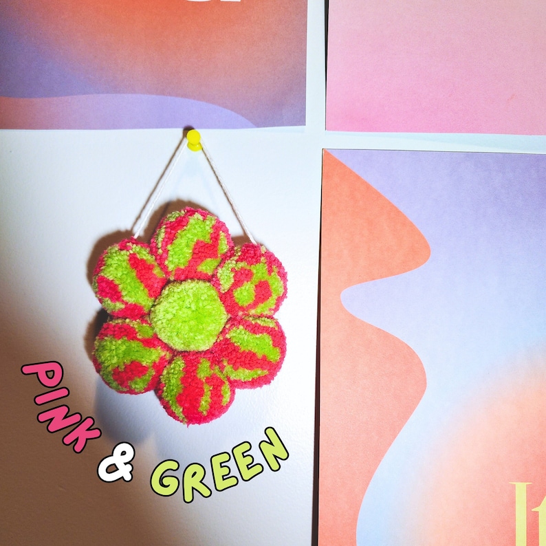 May include: A handmade, flower-shaped wall hanging made of pink and green yarn. The flower is attached to a white wall with a yellow push pin. The text "PINK & GREEN" is written in a playful font below the flower.