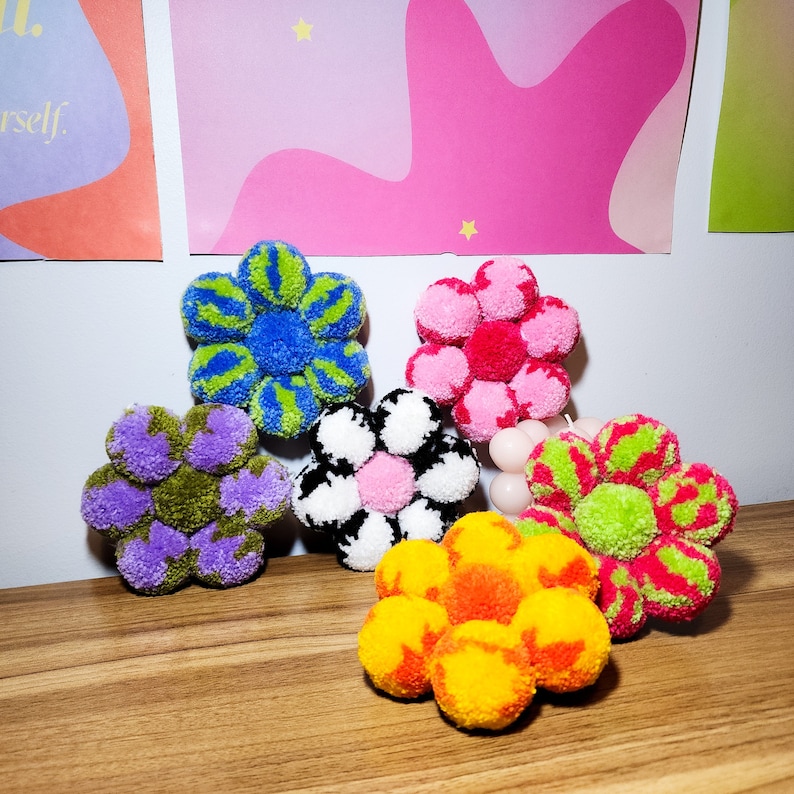 May include: A collection of handmade pom-pom flowers in various colors, including blue, pink, purple, black and white, and orange. Each flower is composed of multiple pom-poms arranged in a floral shape. The flowers are displayed on a wooden surface.