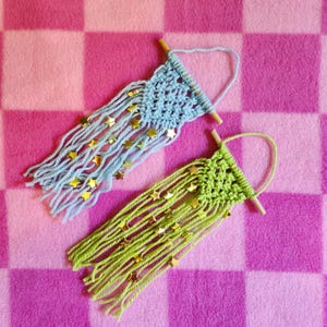 May include: Two macrame wall hangings with gold star accents. One is light blue and the other is green.