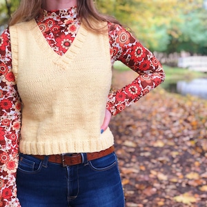 May include: A pale yellow knitted vest with a V-neck and a brown leather belt. The vest is worn over a red and brown floral patterned long-sleeved shirt.