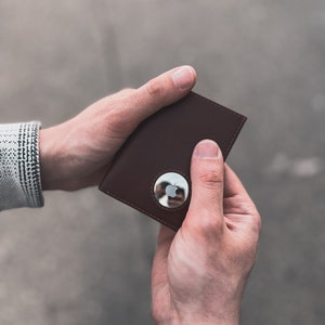 May include: A brown leather wallet with a silver Apple AirTag attached. The wallet is rectangular and held between two hands. The background is a blurred gray. The AirTag is circular with the Apple logo.