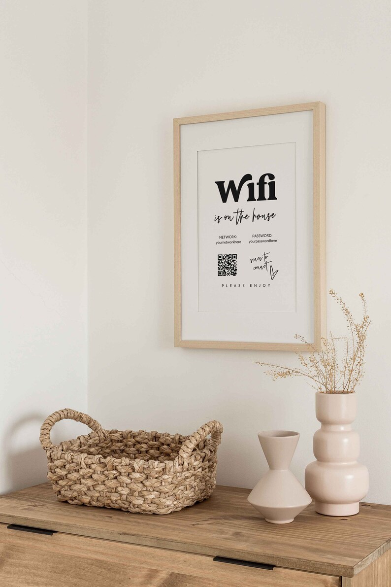May include: A framed sign with the text "Wifi is on the house" and a QR code. The sign also includes the words "Network: yournetworkhere" and "Password: yourpasswordhere". Below the QR code is the text "scan to connect" and a heart symbol. The sign is on a white wall with a wooden frame. There is a wicker basket and two ceramic vases on a wooden table below the sign.