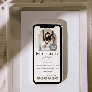 May include: A white and gray mobile phone mockup with a business card displayed on the screen. The card features a photo of a person holding a camera, the text "Marie Louise Photography", a phone number, email address, website address, social media handle, and icons for Instagram, LinkedIn, Pinterest, Facebook, TikTok, and Twitter. The text "Let's Be Social" is also displayed on the card.