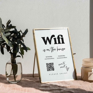 May include: A gold framed sign with the text "Wifi is on the house" and a QR code. The sign is on a stand and there is a vase with greenery and a candle on a pink surface.