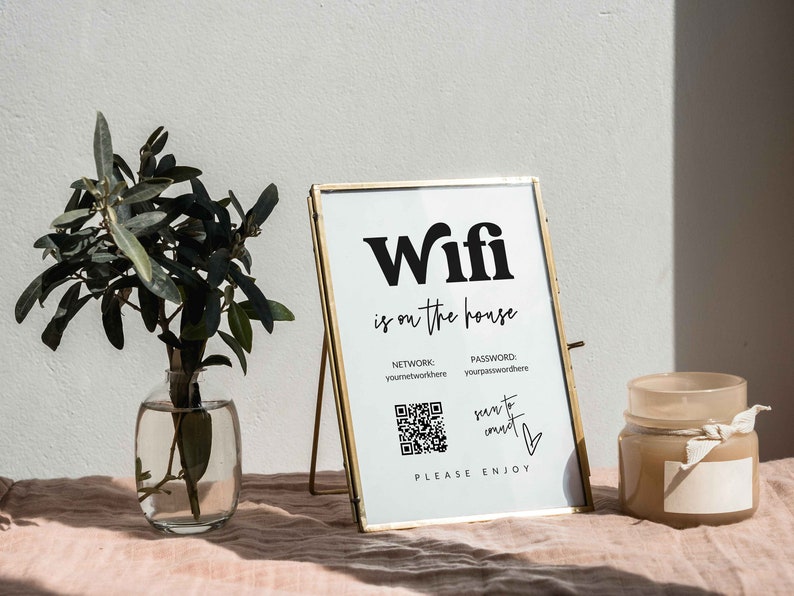 May include: A gold framed sign with the text "Wifi is on the house" and a QR code. The sign is on a stand and there is a vase with greenery and a candle on a pink surface.