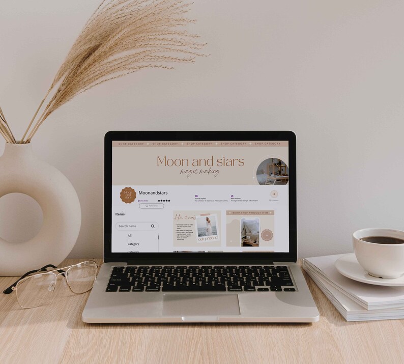 K&ouml;nnte beinhalten: Ein Laptop-Computer zeigt eine Website f&uuml;r einen Shop namens "Moon and Stars" mit dem Slogan "magic making". Die Website verf&uuml;gt &uuml;ber eine Suchleiste, ein Kategoriemen&uuml; und ein Raster mit Produktbildern. Der Laptop steht auf einem Holztisch mit einer Tasse Kaffee und einem Stapel B&uuml;chern.