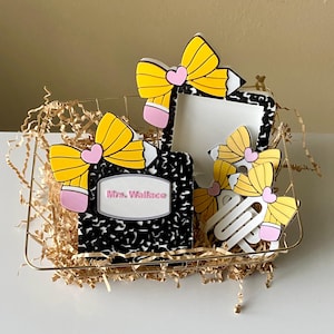 May include: A gold wire basket filled with decorative items. The basket contains a notebook-shaped item with the text "Mrs. Wallace", a white mug, and several pencil-shaped decorations with yellow, pink, and black accents. The basket is filled with shredded paper.