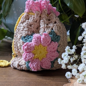 May include: A crocheted drawstring bag with a pink flower design. The bag is made with beige yarn and has a pink and yellow flower on the front.