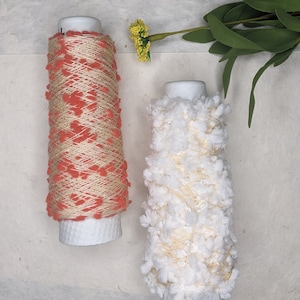 May include: Two spools of yarn, one with a white fluffy texture and the other with a peach and gold textured pattern.