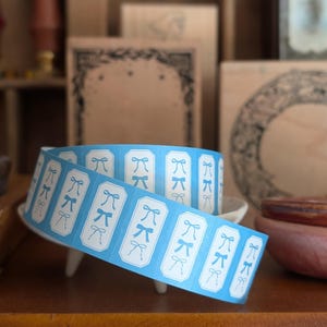 May include: A roll of light blue stickers with white bow illustrations. The stickers are rectangular with rounded corners and are arranged in a continuous strip. The roll is resting on a small white dish.
