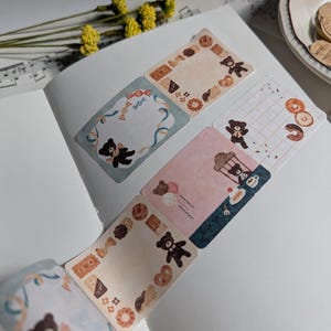 May include: Five decorative stickers featuring a cartoon bear with a variety of backgrounds and designs. The stickers include a blue and white frame with a ribbon, a pink and blue background with a bear holding a balloon, a brown and white checkered background with a bear holding a cup of tea, a brown and white background with a bear holding a cookie, and a brown and white background with a bear holding a cookie.