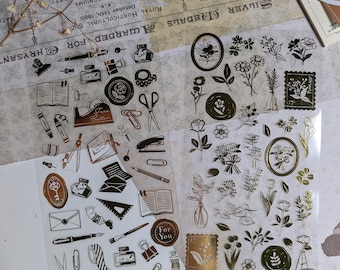 May include: Two sheets of clear stickers with gold and black illustrations. One sheet features stationery items like pens, scissors, and books. The other sheet has floral designs, including vases and stamps. The text "For You" is visible on one sticker.