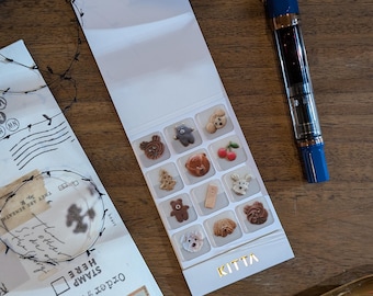 May include: A collection of decorative stickers featuring various animal and food designs, including bears, cherries, and dogs. The stickers are arranged on a white card with the word "KITTA" in gold. A blue fountain pen is also visible.