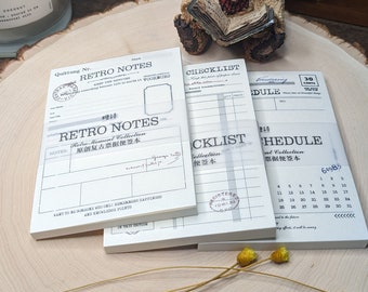 May include: A stack of retro-style notepads with printed text and graphics. The pads feature designs like "Retro Notes," "Checklist," and "Schedule." A small teddy bear figurine sits atop the stack, and dried yellow flowers are in the foreground.