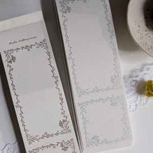 May include: Two stacks of blank notepads with floral borders. The top notepad has a light blue floral border and the bottom notepad has a brown floral border. Both notepads have the text "coda letterpress" printed on the front.