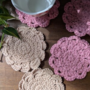 May include: Four crocheted coasters, two in a light beige color and two in a dusty rose color. The coasters are round with a scalloped edge and a textured design.