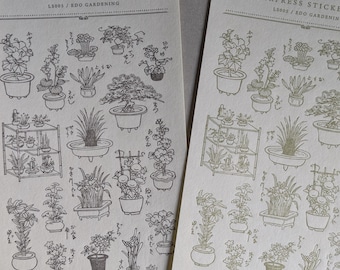 May include: Two sheets of letterpress stickers featuring illustrations of potted plants and flowers. The stickers are gold on a cream background. The text on the sheets reads "Letterpress Sticker Sheet" and "LS005/Edo Gardening". The bottom of the sheets reads "WACCA x OEDA LETTERPRESS".