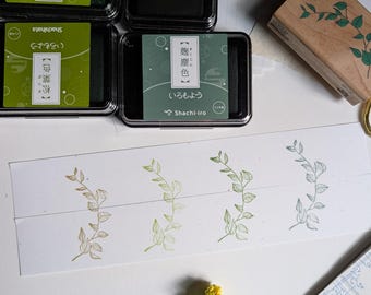 May include: An overhead shot features four ink pads in green, brown, and gold tones, with Japanese text. A wooden stamp with a leaf design and stamped leaf patterns in various colors are also visible. Yellow flowers and stationery complete the scene.