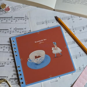 May include: A spiral-bound notebook with a red cover featuring a whimsical illustration of a teacup with a cat inside and a girl in a dress. The notebook is open on a surface with sheet music and a pencil.
