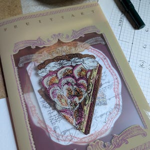 May include: A decorative piece featuring a slice of fruit tart with a cream topping. The tart is set within a clear frame, with the word "FRUIT TART" at the top. The design includes a decorative border and is set on a light-colored background.