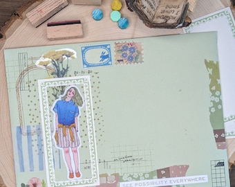 May include: A green envelope with a variety of decorative stickers and stamps. The envelope has a white border and the text "SEE POSSIBILITY EVERYWHERE" printed on the bottom. The envelope is on a wooden surface.